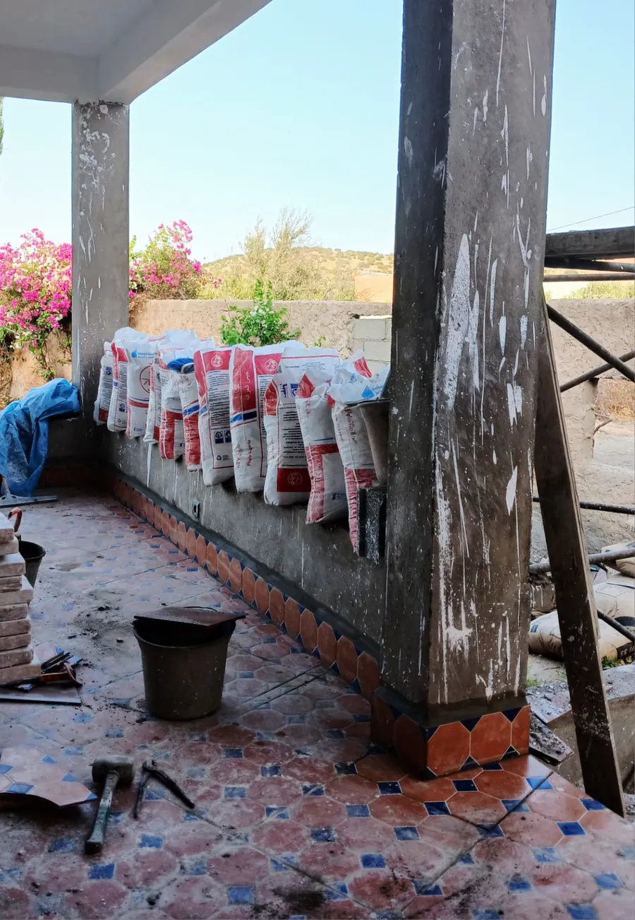 Des carreaux empilés et des sacs alignés le long d'un muret, sous un auvent au sol carrelé.