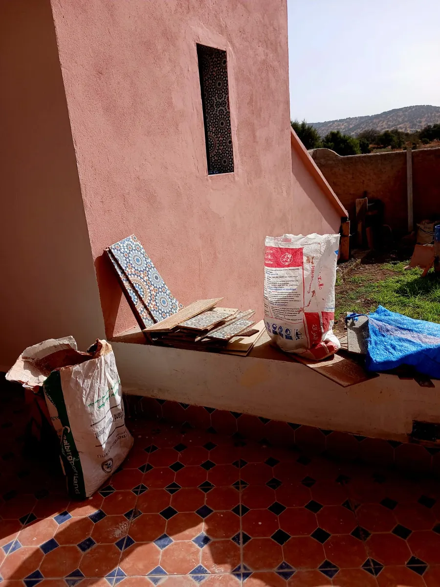 Des zelliges appuyés contre un muret rose, à côté de sacs de ciment, sur un sol carrelé.