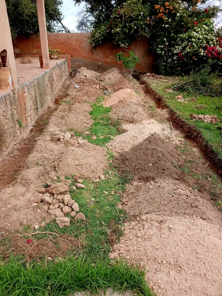 Une tranchée creusée le long d'un mur de terre, entre une pelouse et des massifs fleuris.